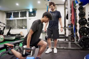 後ろ足をベンチに乗せダンベルで片脚スクワットをする男性と、横で見守るトレーナー。体の土台づくり
