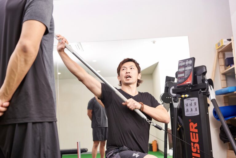 ケーブルマシンでバーを斜めに引き、体をひねる動きを鍛えるトレーニングと、見守るトレーナー