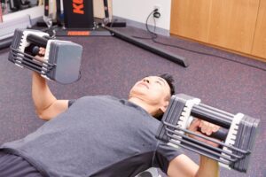 ベンチで両手のダンベルを押し上げる男性と奥のトレーニングマシン。筋トレ初心者の負荷の決め方