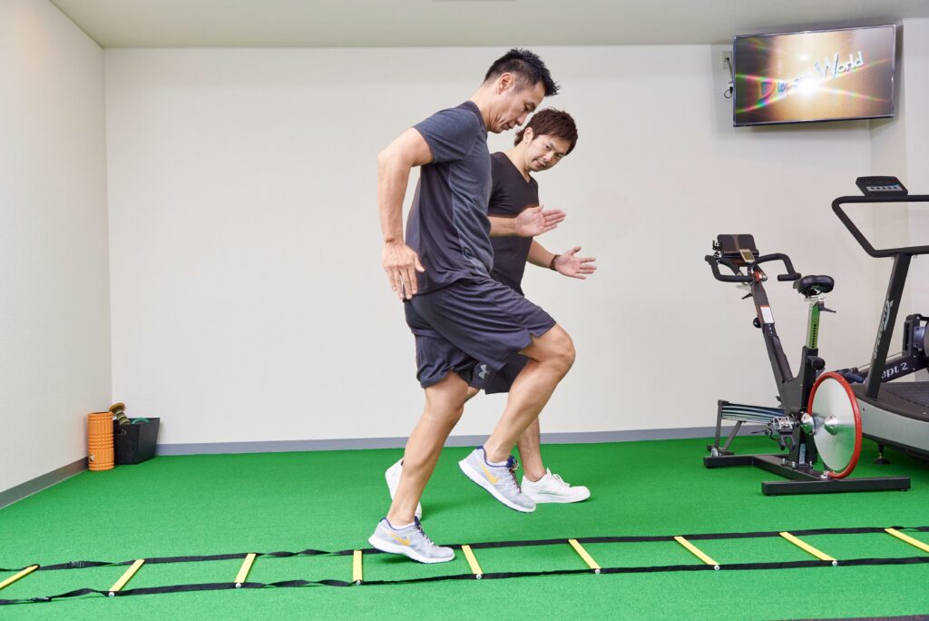 人工芝に置いたラダーでステップ練習をする男性と、手振りを交えて見守るトレーナー。動ける身体づくり