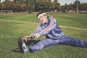 サッカー場の芝生で顔をしかめながらつま先へ手を伸ばすジャージ姿の人。昼休みの短い運動